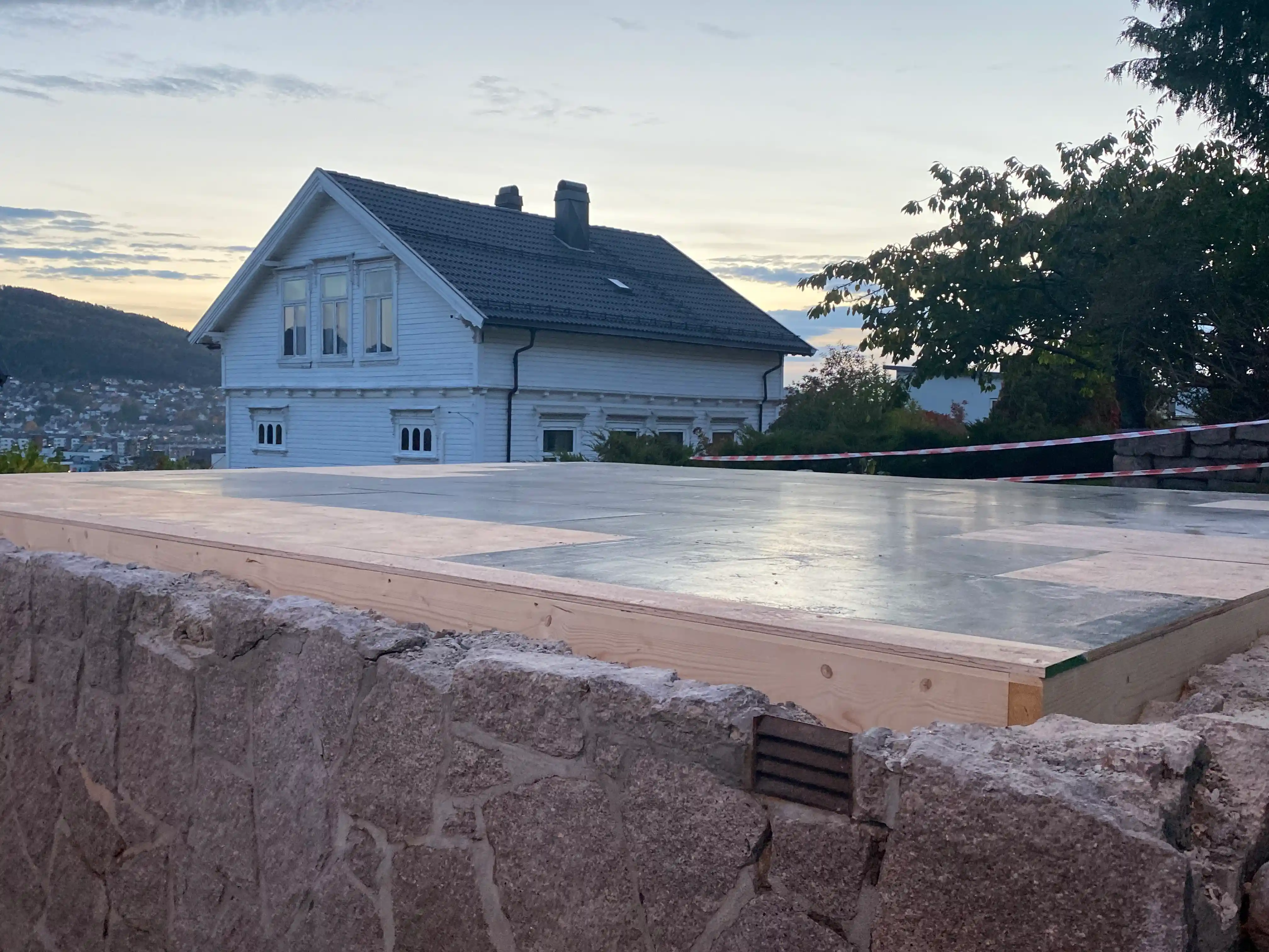 Byggeplass med grunnmur og hvitt hus i bakgrunnen.